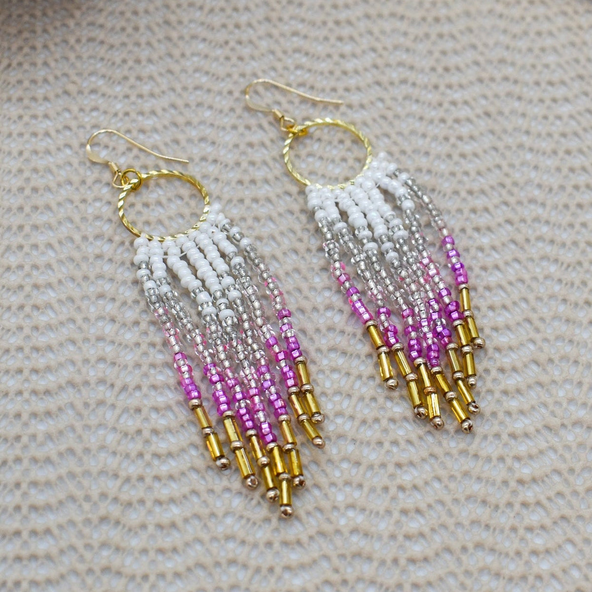Beaded earrings with white, pink, and gold beads on a textured surface.