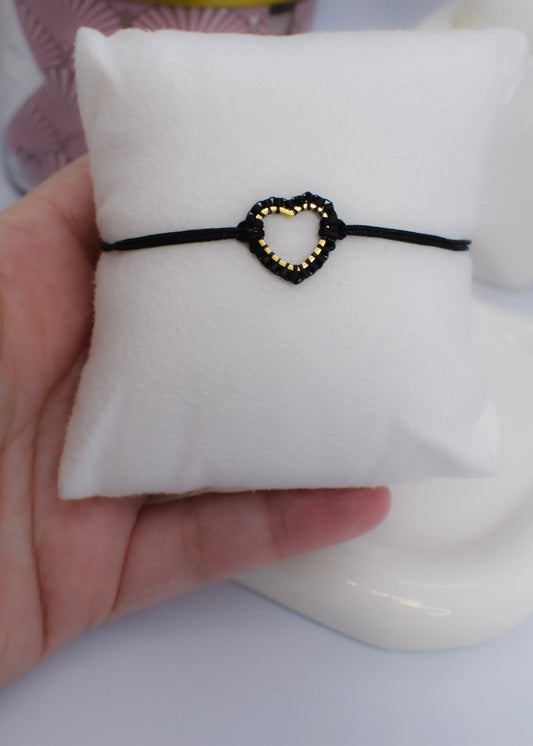 Black bracelet with a heart-shaped Miyuki beaded charm on a white cushion, held by a hand.