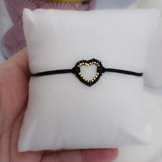 Black bracelet with a heart-shaped Miyuki beaded charm on a white cushion, held by a hand.