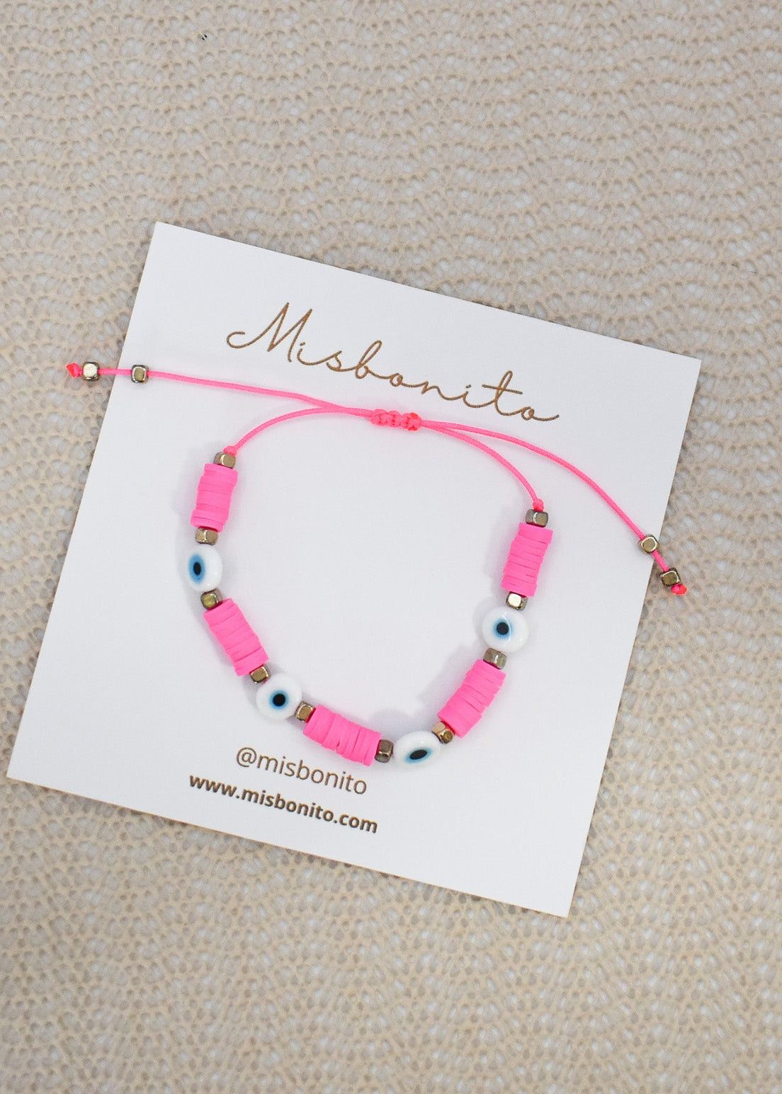 A pink beaded bracelet with evil eye beads displayed on a white card labeled Misbonita, set against a textured beige background.