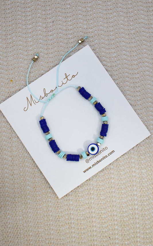 A beaded bracelet with royal blue and light blue beads, a central evil eye charm, and small gold spacer beads, displayed on a white Misshanti brand card over a beige textured fabric background.