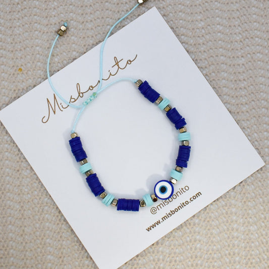 A beaded bracelet with royal blue and light blue beads, a central evil eye charm, and small gold spacer beads, displayed on a white Misshanti brand card over a beige textured fabric background.