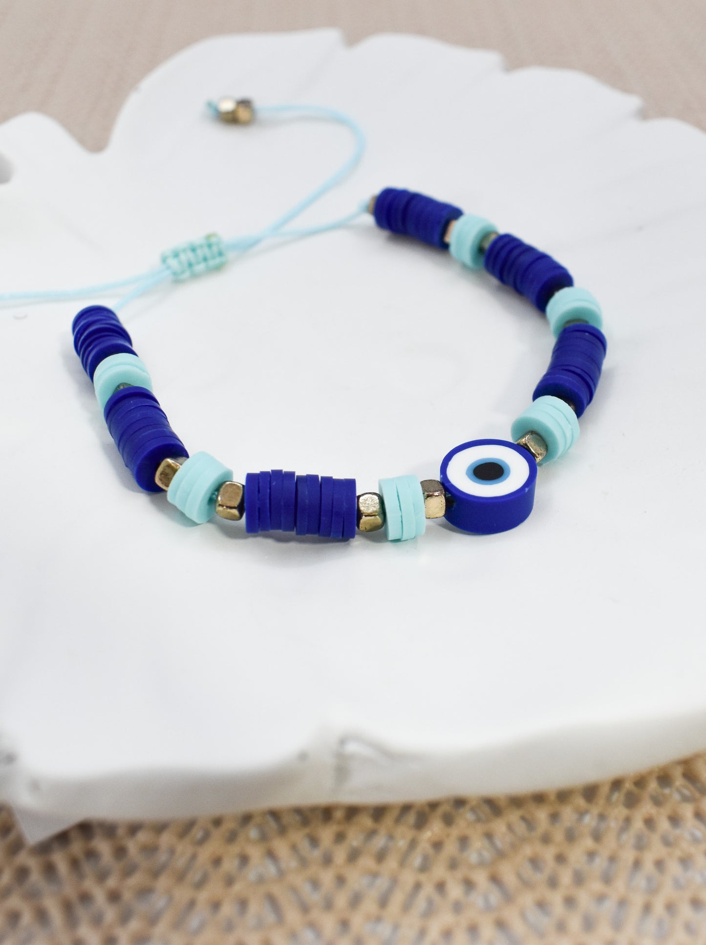 A beaded bracelet with dark blue and light blue cylindrical beads, a blue evil eye charm, and small gold spacer beads, displayed on a white decorative tray.