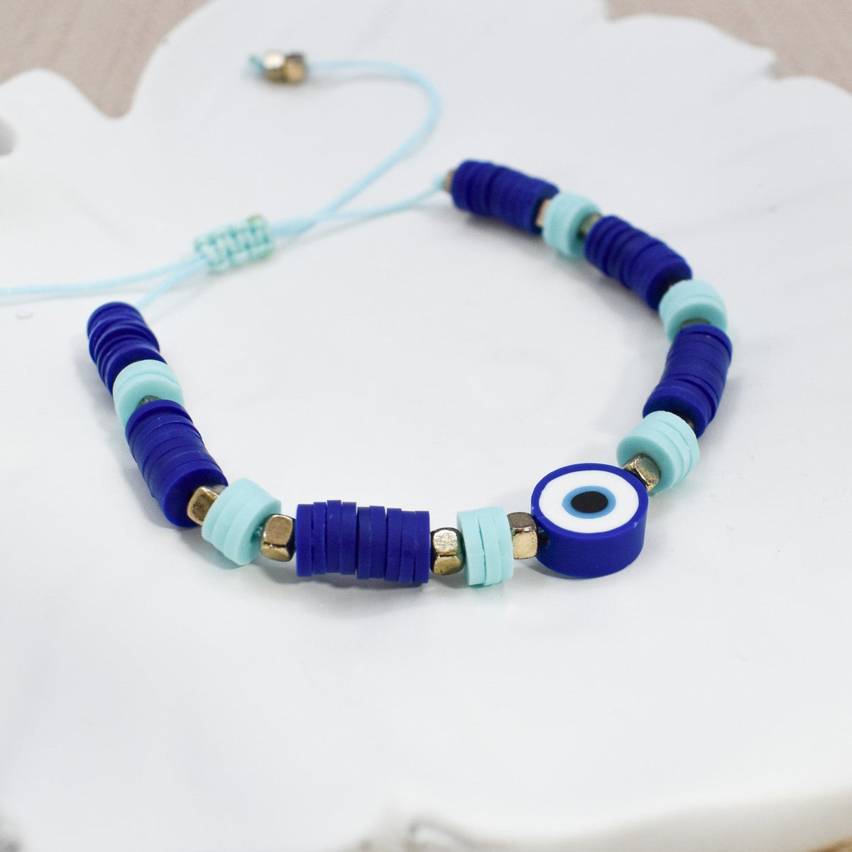 A beaded bracelet with dark blue and light blue cylindrical beads, a blue evil eye charm, and small gold spacer beads, displayed on a white decorative tray.