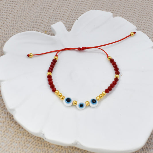 A red cord bracelet with small red and gold beads, featuring three white evil eye charms with blue details, displayed on a white ceramic leaf-shaped dish over a beige textured fabric background.