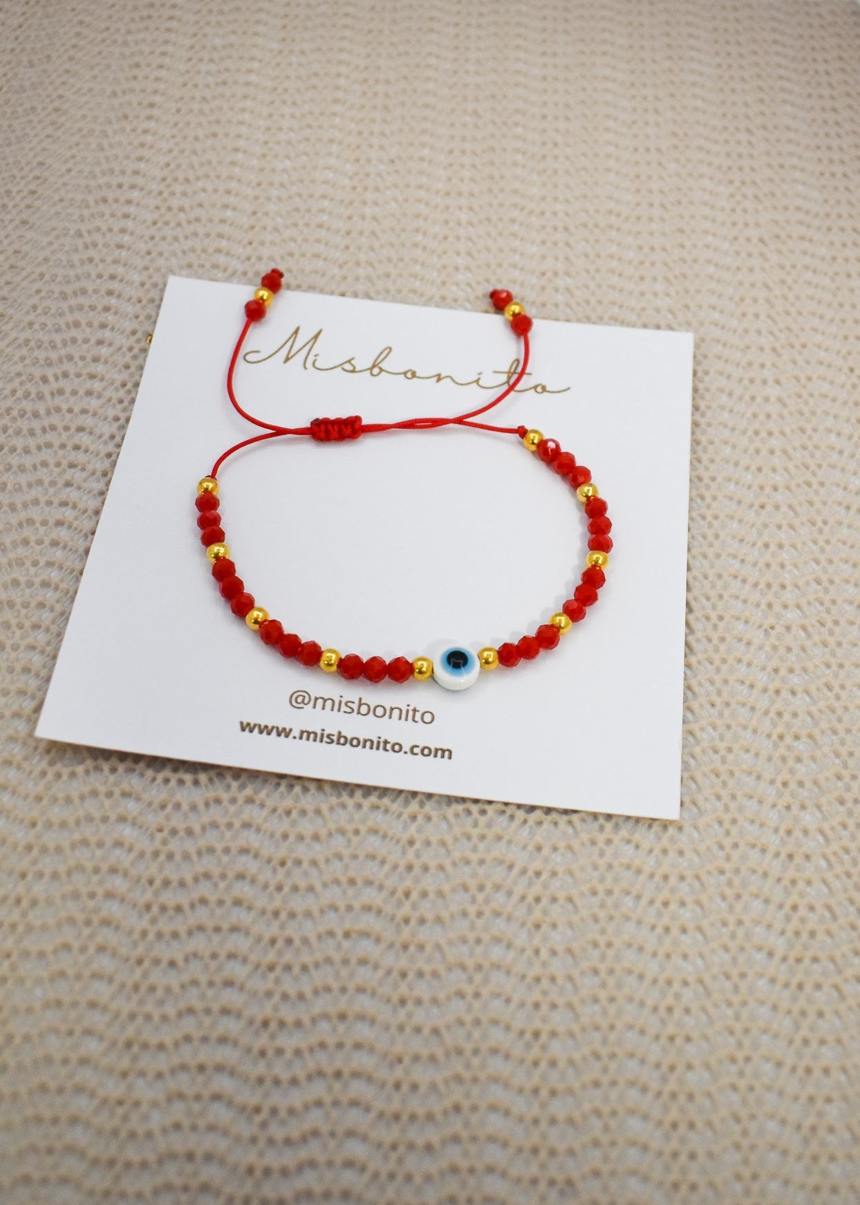 A red beaded cord bracelet with a blue and white evil eye charm, displayed on a white branded card over a beige textured fabric background.