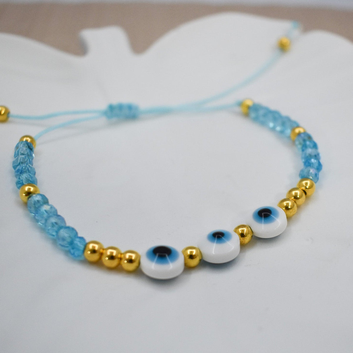 A beaded bracelet with light blue beads, shiny gold spacer beads, and three white evil eye beads, displayed on a white surface.