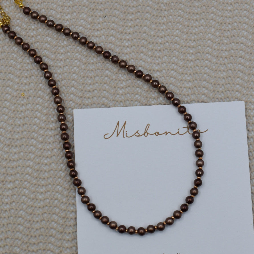 A dark brown beaded necklace with gold-toned hardware, displayed on a white brand card against a textured beige woven background.