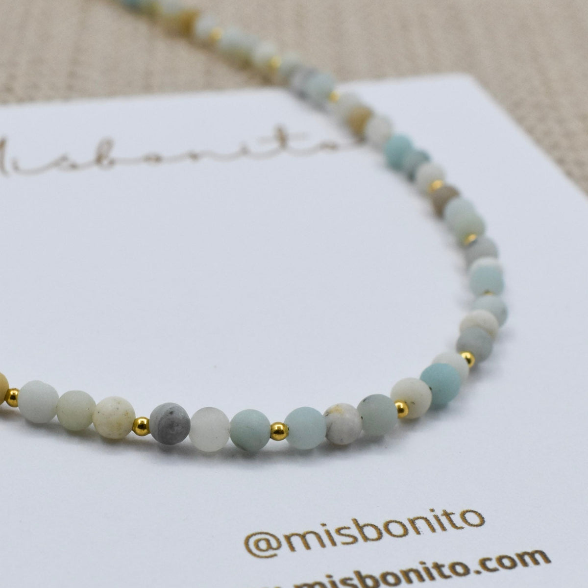 zoomed mini Amazonite beads bracelet with light blue-green beads and gold-tone details, displayed on a white misbonito card on a beige textured fabric background.