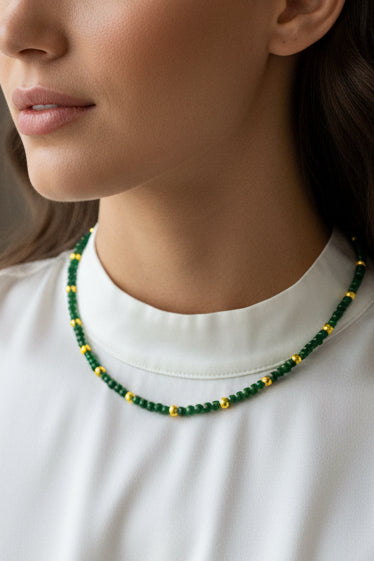 A beaded necklace with dark green and gold beads displayed on a white card labeled Misbonito, set against a textured beige fabric background.