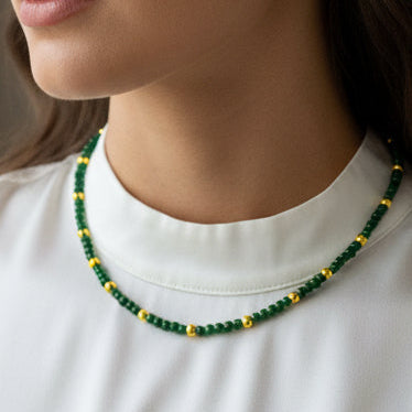 A beaded necklace with dark green and gold beads displayed on a white card labeled Misbonito, set against a textured beige fabric background.