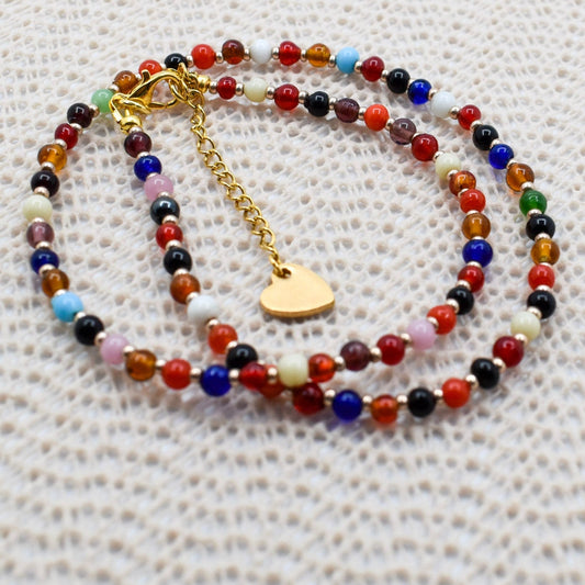 A beaded necklace with multicolor round beads and a gold heart charm, displayed on a textured white fabric background.