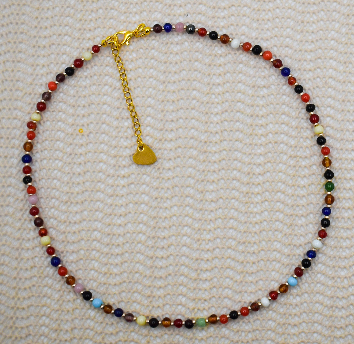 A beaded necklace with multicolor round beads and a gold heart charm, displayed on a textured cream woven fabric background.