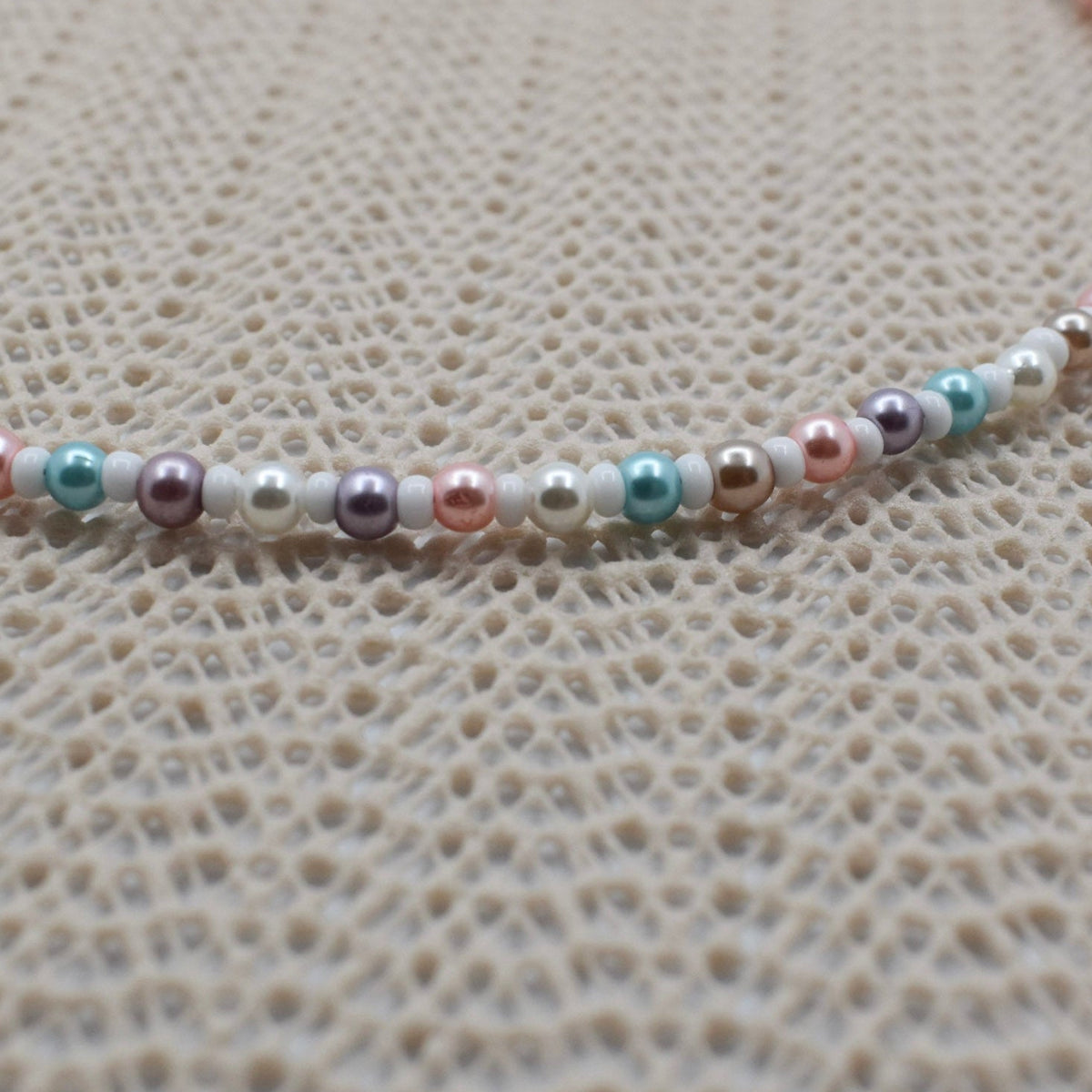 A close-up of a beaded necklace featuring small pearls in pastel pink, mint green, lavender, and white, laid out on a textured beige woven surface.