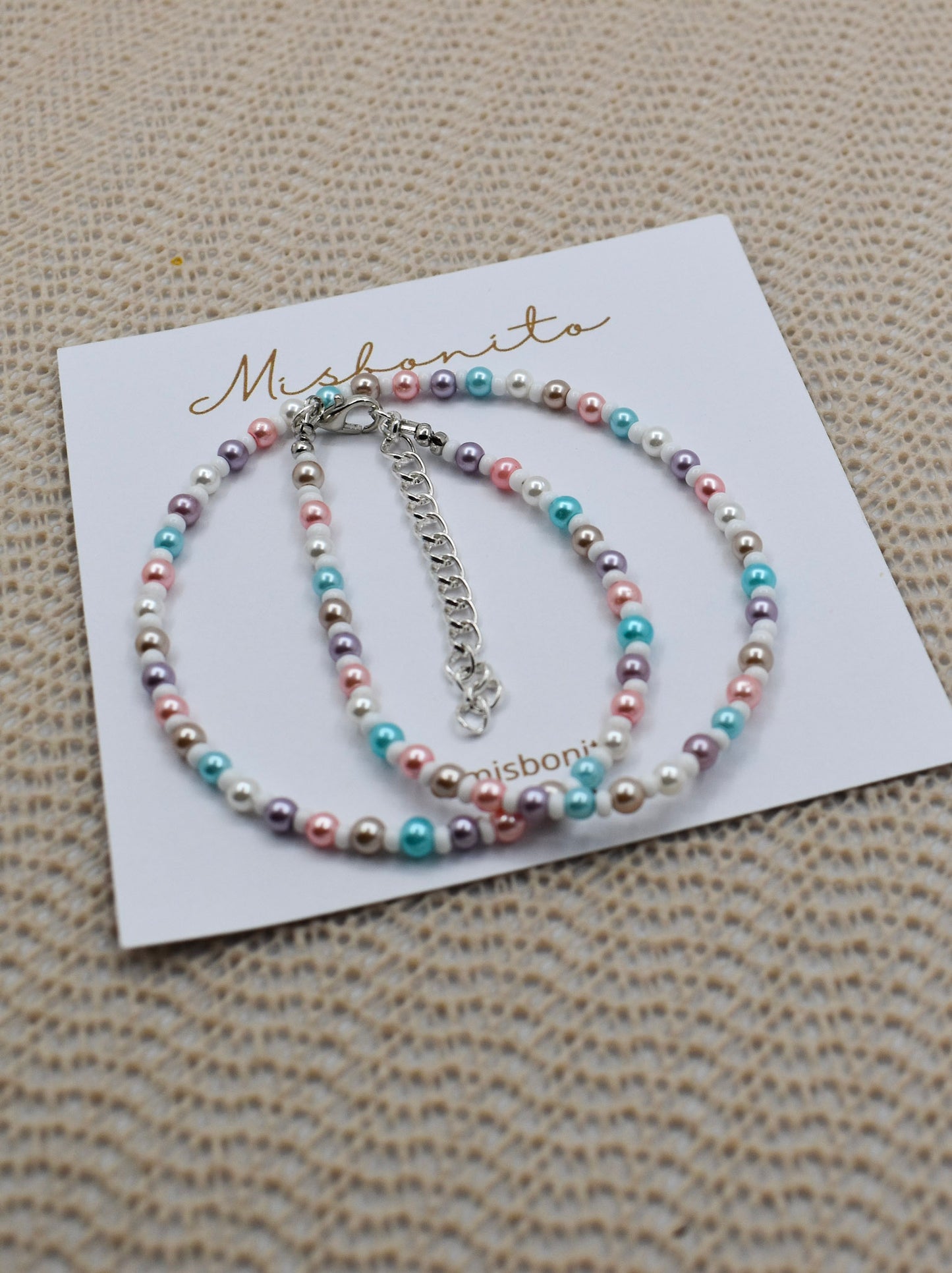 A beaded bracelet with pastel pink, blue, purple, and white pearls on a silver chain, displayed on a white card printed with the brand name 'Misbonita' on a textured beige woven background.