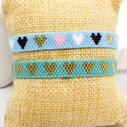 Two handcrafted Miyuki bracelets with a multiheart pattern, one in blue and the other in a lighter shade, displayed on a beige small display pillow.