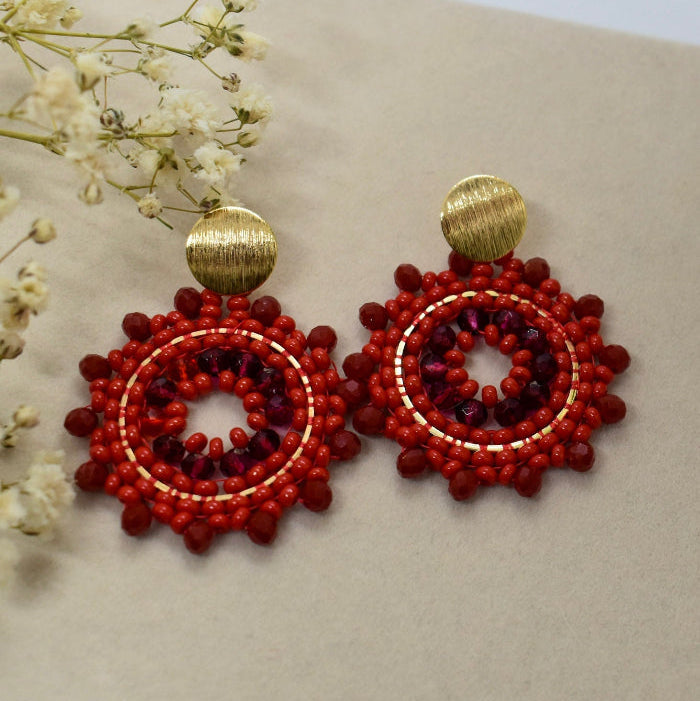 Red beaded earrings with gold accents on a beige surface with white flowers.