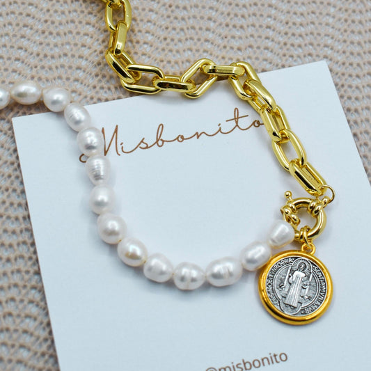 A gold chain bracelet with a white pearl strand and a Saint Benedict medallion charm, displayed on a white Misbonito brand card over a textured beige fabric background.