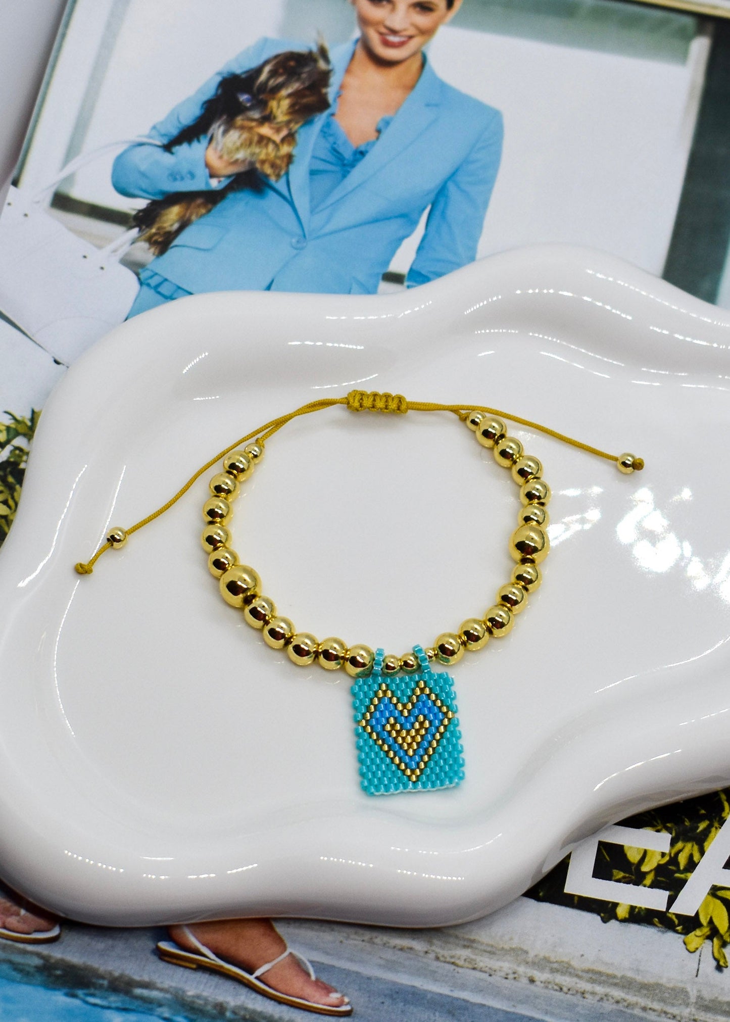 Gold beaded bracelet with a blue heart charm on a white dish.