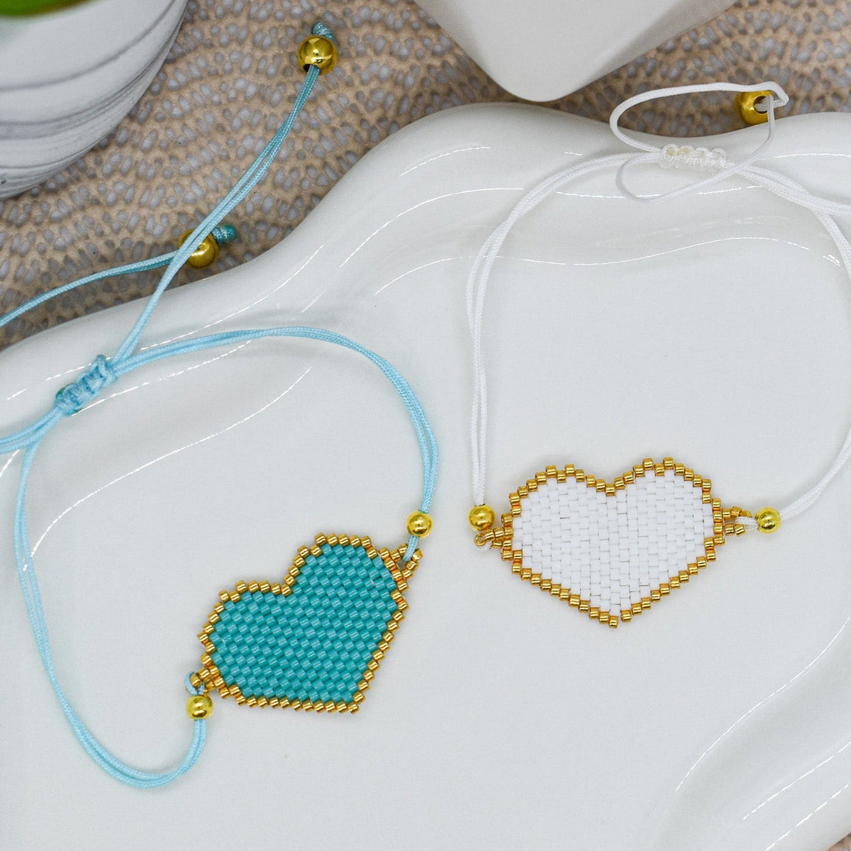 Two heart-shaped beaded bracelets on a white dish with a plant in the background.