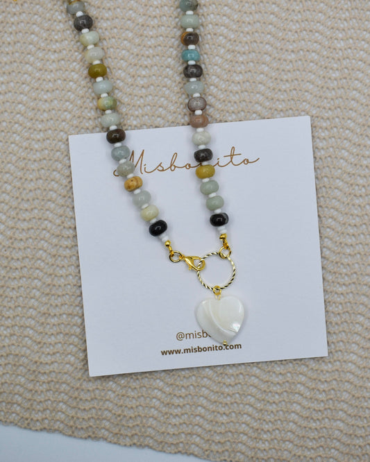A beaded amazonite necklace with a white heart-shaped pendant displayed on a white card labeled Misbonto, placed on a textured beige woven surface.