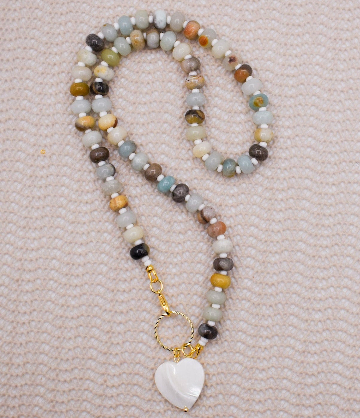 A beaded necklace with amazonite necklace in shades of gray, brown, yellow, and blue, featuring a gold-tone clasp and a white heart-shaped pendant, laid flat on a textured light pink fabric background.