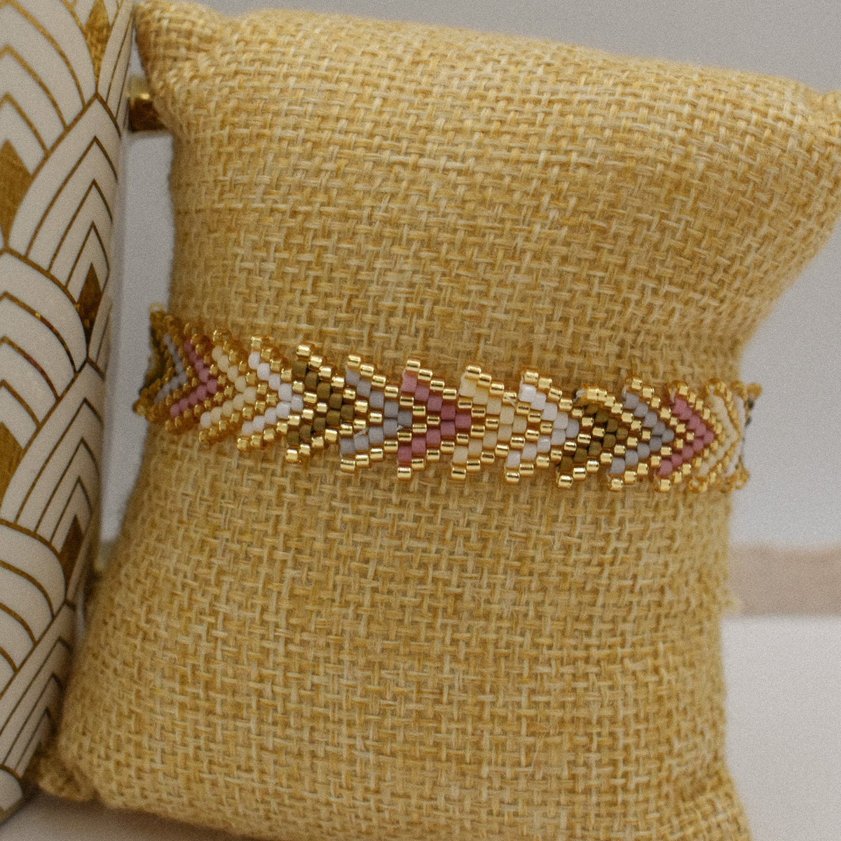A gold beaded chevron-patterned bracelet displayed on a yellow burlap pillow, with a decorative ceramic vase partially visible on the left.