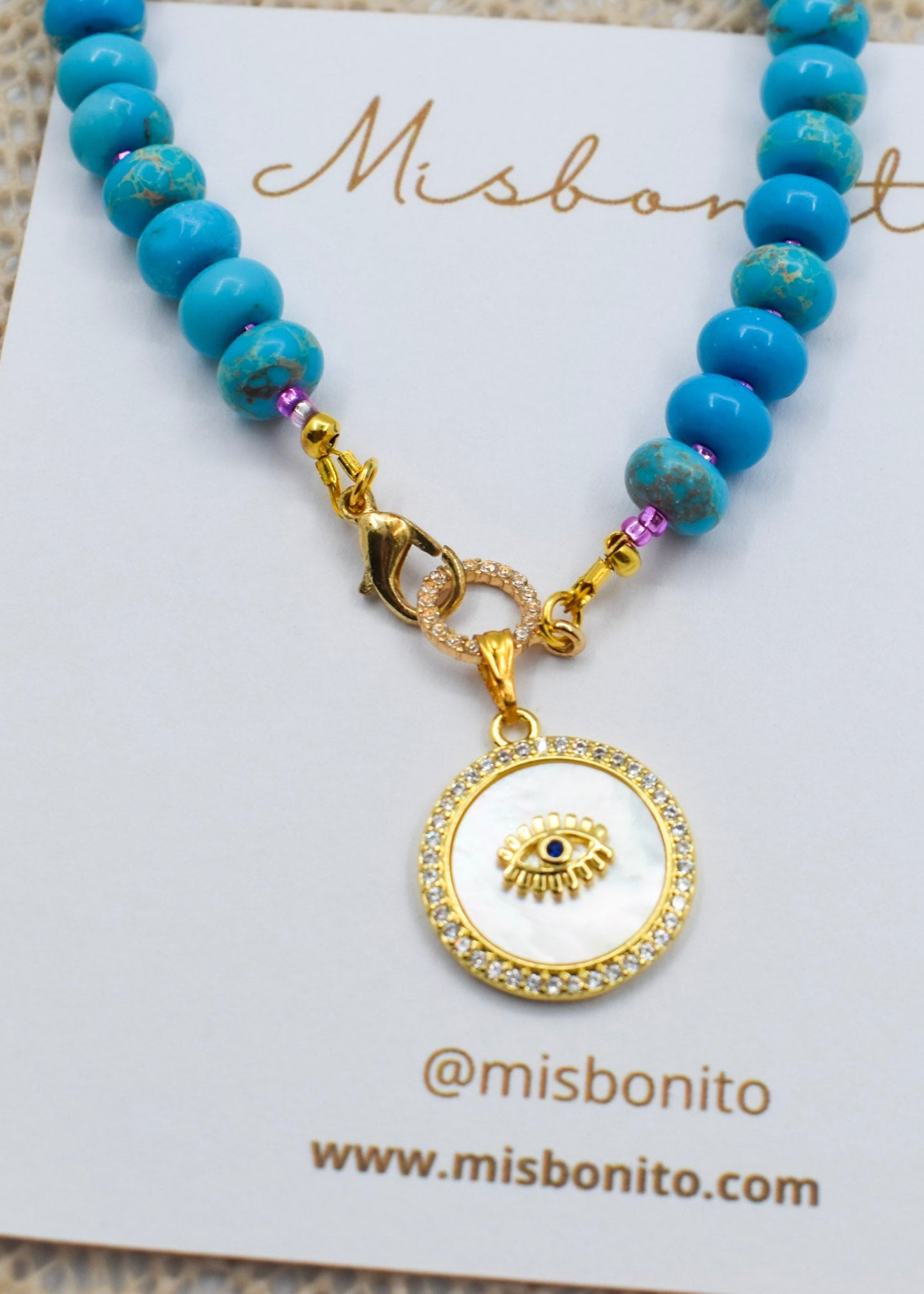 A jasper and gold beaded necklace with a round white and gold pendant featuring an eye design, displayed on a white Misbonito card.
