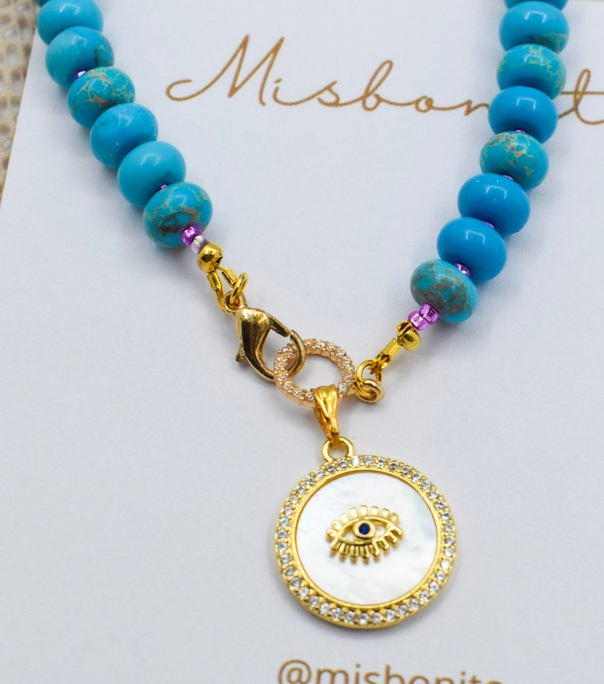 A blue jasper ande evil eye gold beaded necklace with a round white and gold pendant featuring a round eye charm, displayed on a white Misbonito card.