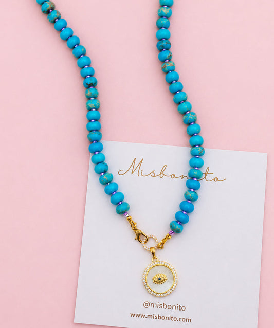 A blue jasper beaded necklace with a gold-toned circular pendant featuring a central eye design, displayed on a white card over a beige textured fabric background.