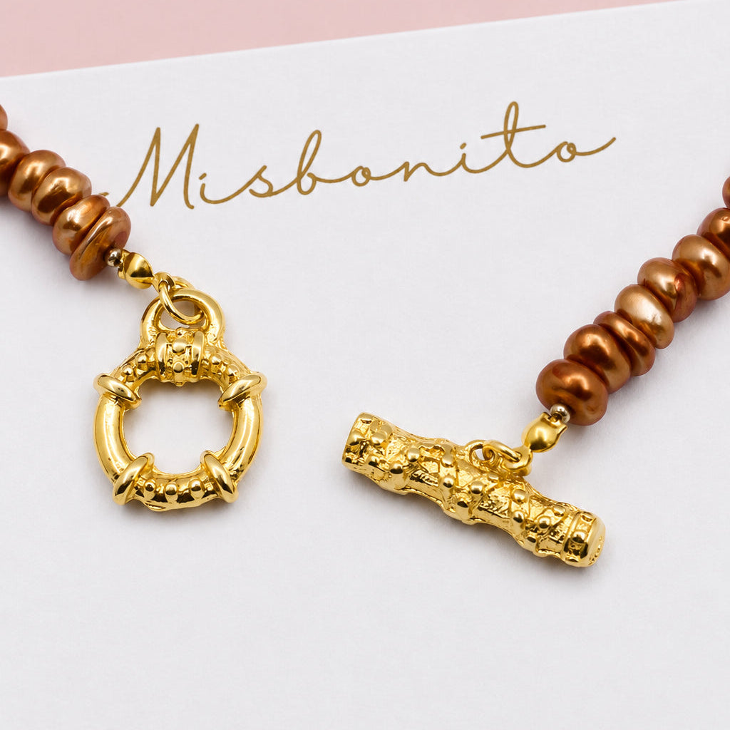 A close-up of a Misbonito beaded necklace with brown beads and gold-toned toggle charms, displayed on a white card with the Misbonito brand name.