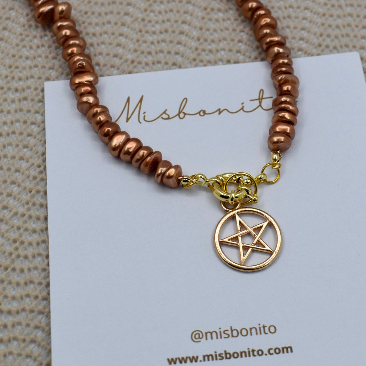 A copper-colored beaded necklace with a gold pentagram pendant, displayed on a white Misbonito brand card over a textured beige fabric background.