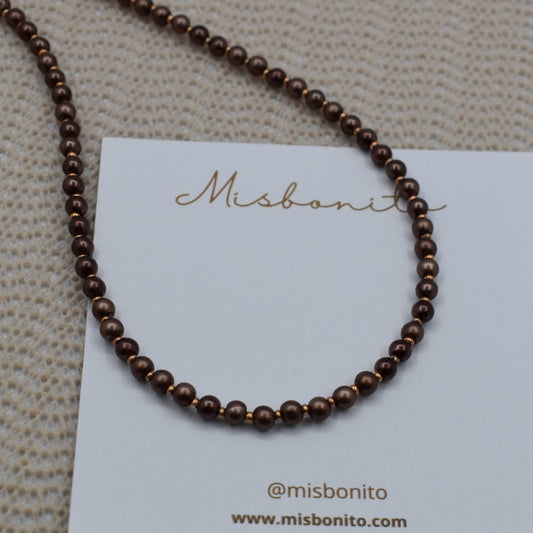 A dark brown beaded necklace displayed on a white Misbonito brand card, set against a textured beige woven background.