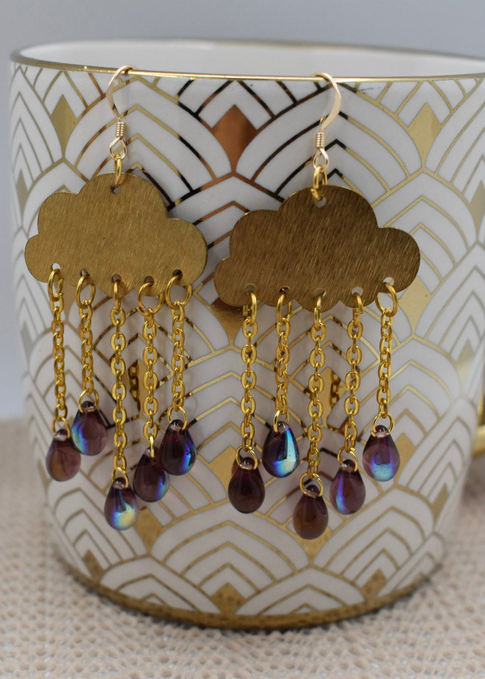A pair of gold cloud-shaped dangle earrings with iridescent teardrop beads, hanging from a white ceramic mug with a gold handle and geometric pattern.