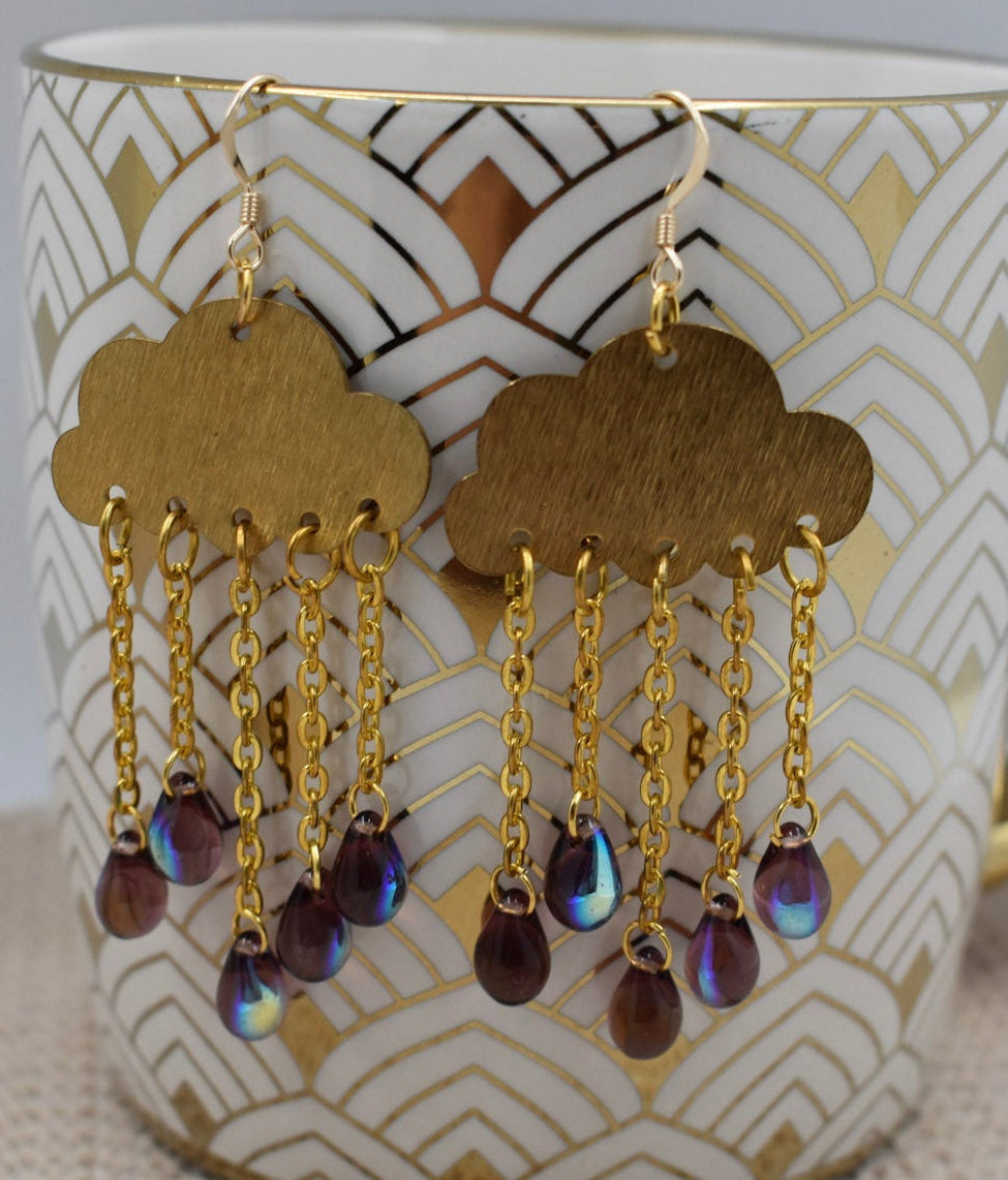 A pair of gold cloud-shaped dangle earrings with iridescent teardrop beads, hanging from a white ceramic mug with a gold handle and geometric pattern.