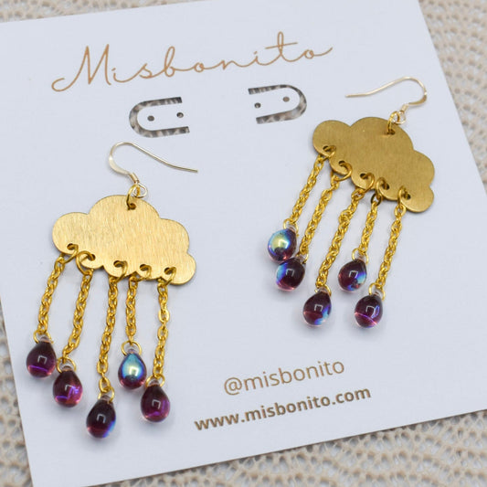 A pair of Misbonito cloud-shaped gold dangle earrings with iridescent purple beads, displayed on a white branded card over a beige knit fabric background.