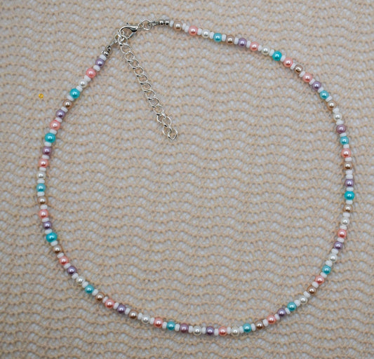 A beaded necklace with small pastel-colored beads in pink, blue, purple, and white, accented by silver-toned spacer beads and a silver clasp with extender chain, laid flat on a textured beige fabric background.