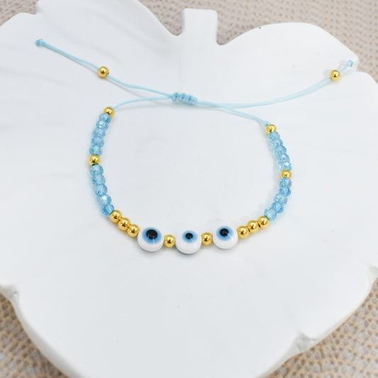 A light blue beaded bracelet with three white evil eye charms and gold spacer beads, displayed on a white ceramic heart-shaped dish over a beige textured fabric background.