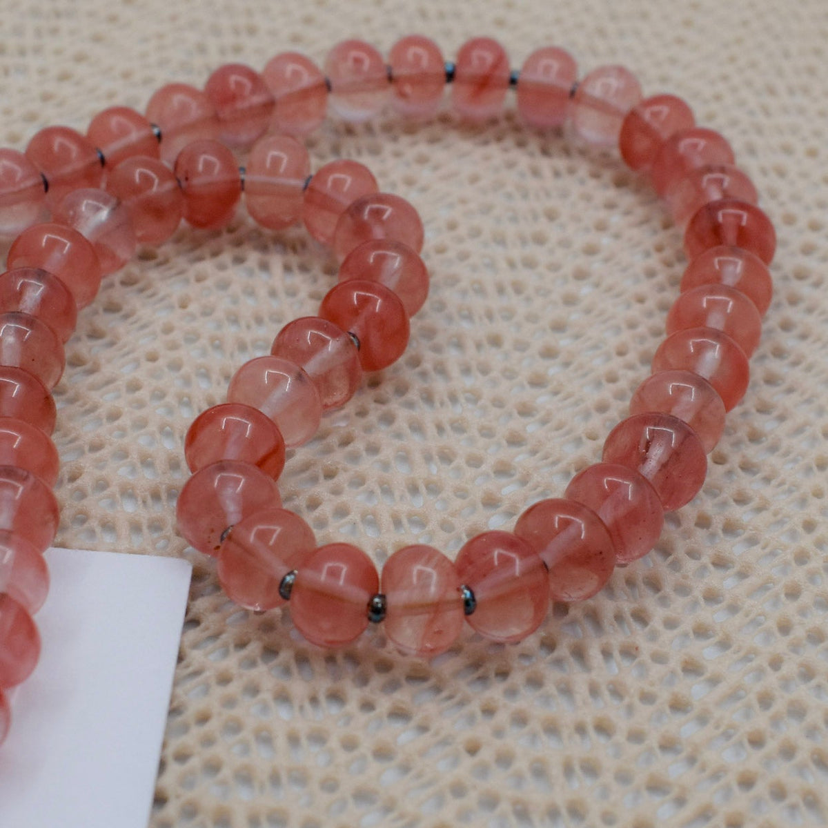 A close up of the strand of translucent pinkish-red round beads with small dark spacer rings, laid on a textured beige woven surface next to a white card.