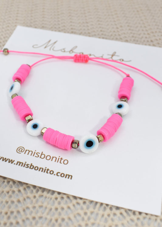 A pink cord bracelet with pink cylindrical beads, white evil eye beads, and small metallic spacer beads, displayed on a white Misbonito card.