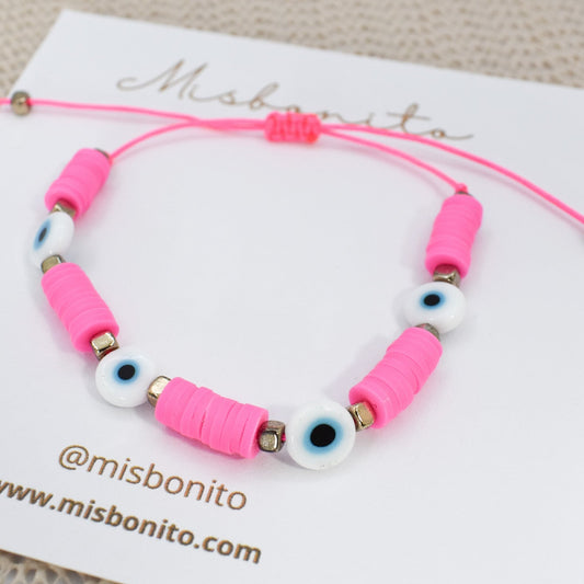A pink cord bracelet with pink cylindrical beads, white evil eye beads, and small metallic spacer beads, displayed on a white Misbonito card.