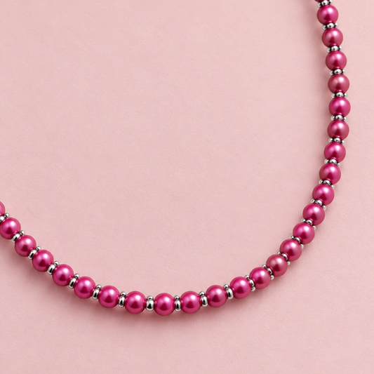 A close-up of a beaded necklace with glossy magenta-pink round beads separated by small metallic spacer rings, draped on a textured beige fabric background.