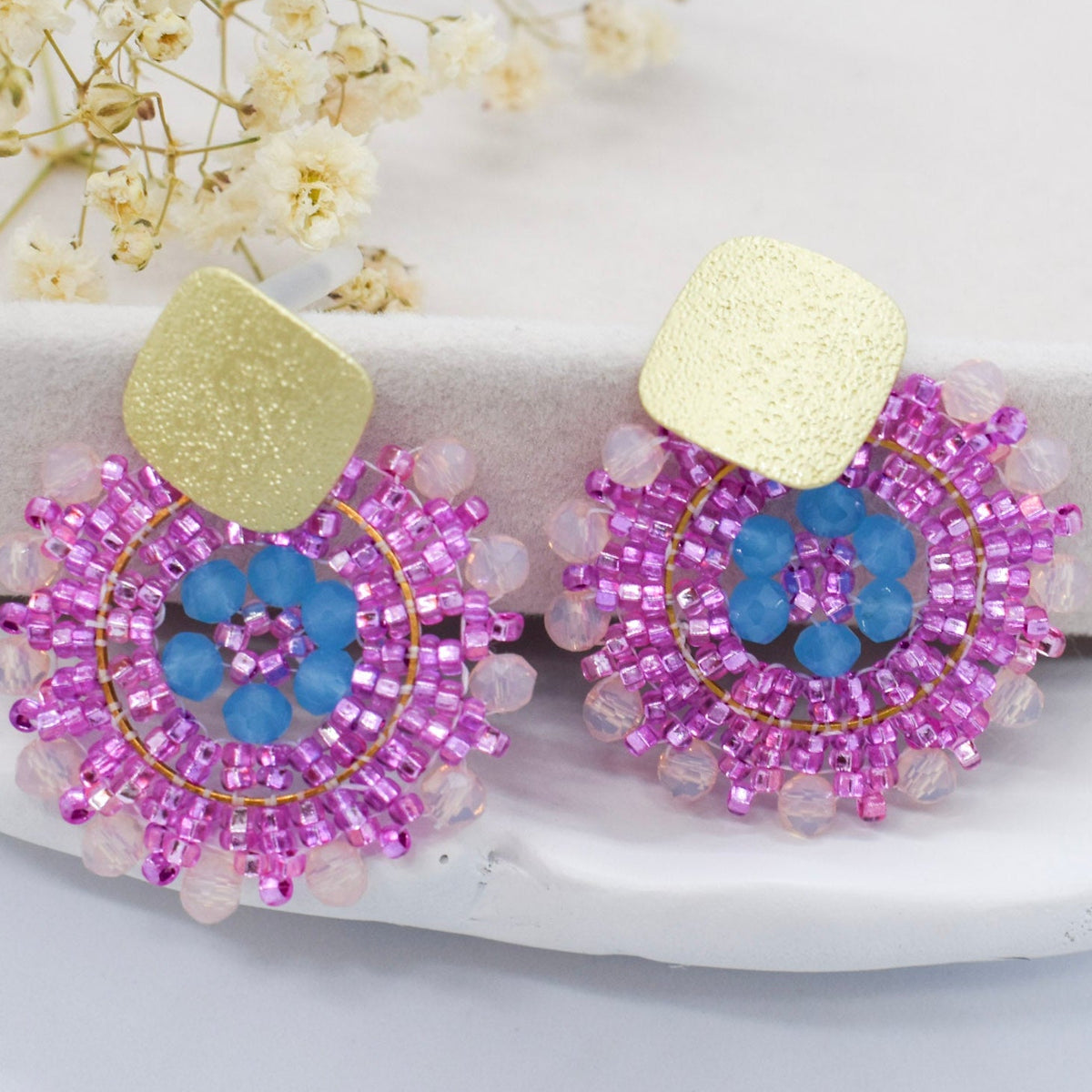 Pair of beaded earrings with pink and blue design on a white surface with flowers in the background