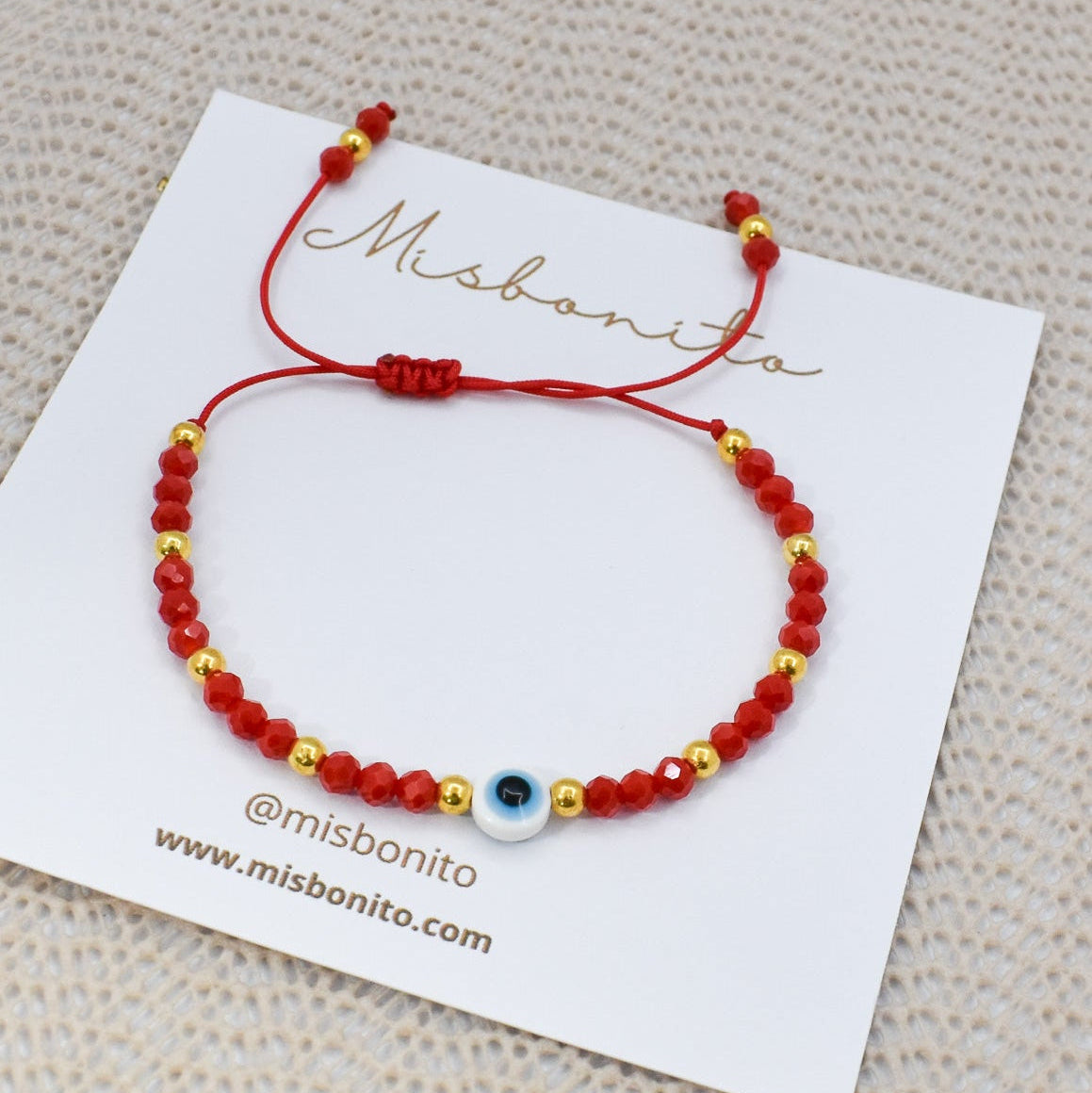 A red beaded bracelet with gold spacer beads and a central blue and white evil eye charm, displayed on a white Misbonito brand card over a textured beige fabric background.