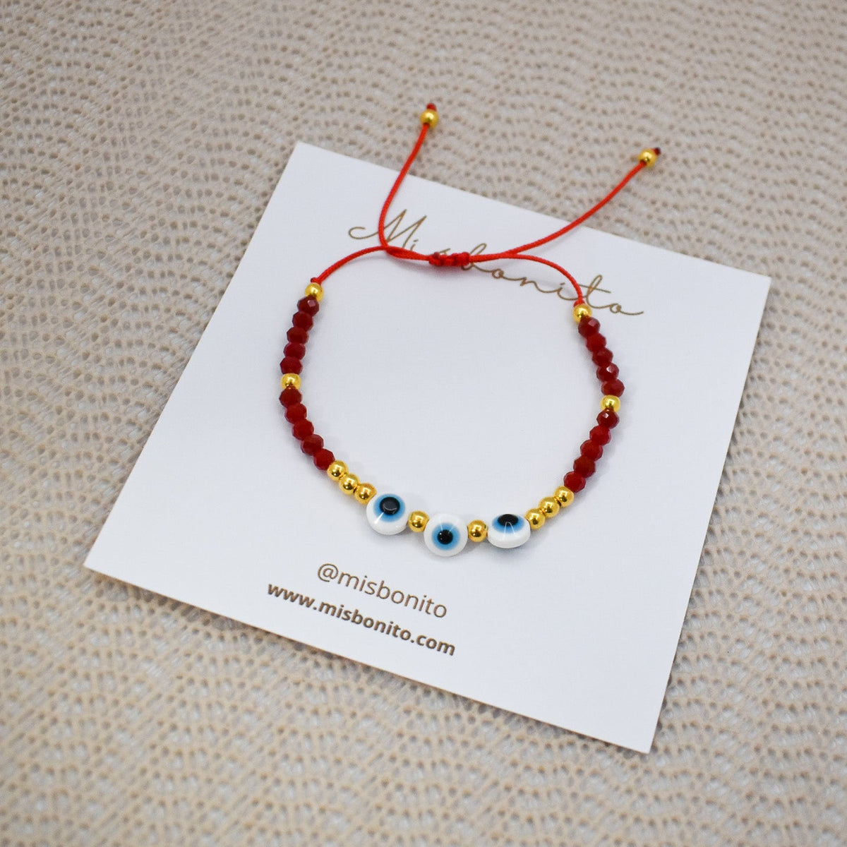 A red cord bracelet with gold spacer beads and three evil eye charms, displayed on a white branded card over a beige textured fabric background.