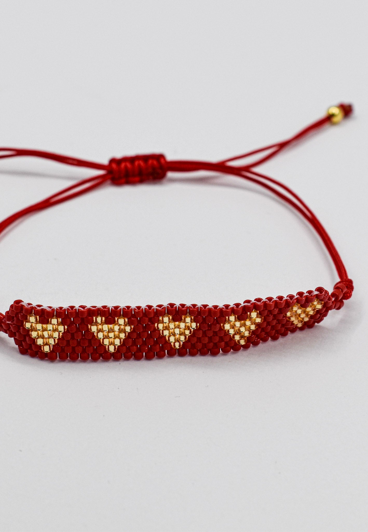 A red beaded bracelet with gold heart patterns and adjustable cord, shown on a light gray background.