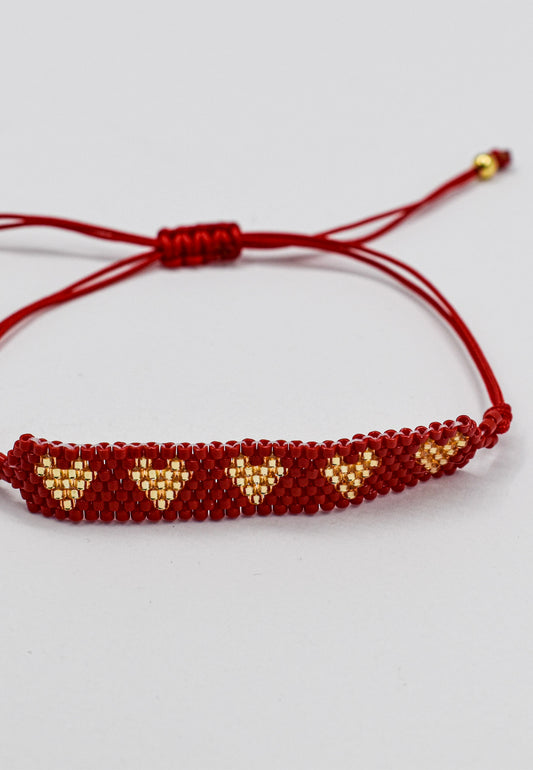 A red beaded bracelet with gold heart patterns and adjustable cord, shown on a light gray background.