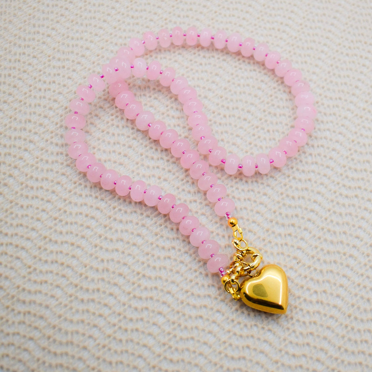 A rose quartz abacus stone necklace with a golden heart charm on a beige textured surface