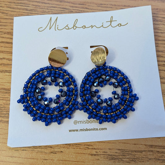A pair of Misbonita beaded drop earrings with gold-tone tops and blue seed beads, displayed on a white brand card on a wooden surface.