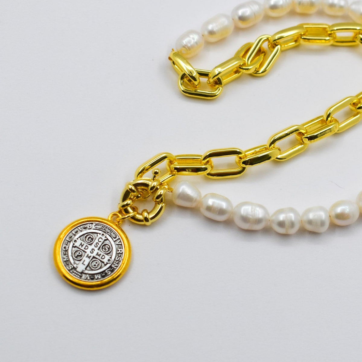 A gold chain necklace with white pearl beads and a round gold Saint Benedict medal pendant, displayed on a light gray background.